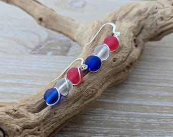 Könnte beinhalten: Handgefertigte Ohrringe mit roten, weißen und blauen Glasperlen, die ein patriotisches Thema aufgreifen. Die Perlen sind auf einem silbernen Draht aufgefädelt, mit einem einfachen Haken-Design. Die Ohrringe werden auf einem Stück Treibholz präsentiert.