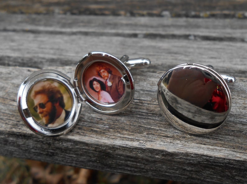 May include: A pair of silver cufflinks with a hinged locket design. One cufflink features a photo of a couple, while the other cufflink is closed and reflective.