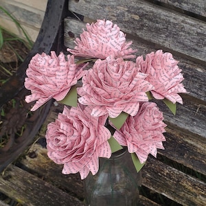 May include: A bouquet of six pink paper flowers made from book pages. The flowers are arranged in a clear glass vase. The flowers are all different sizes and shapes, and they are all made from different pages of a book. The flowers are all facing the camera.