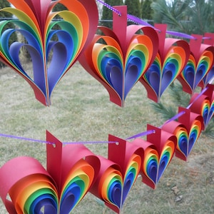May include: A rainbow paper heart garland with red, orange, yellow, green, blue, indigo, and violet paper hearts. The hearts are strung together with purple string.