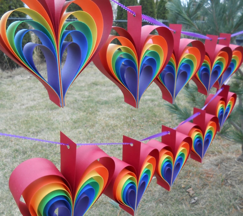 May include: A rainbow paper heart garland with red, orange, yellow, green, blue, indigo, and violet paper hearts. The hearts are strung together with purple string.