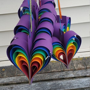 May include: A set of three rainbow heart-shaped paper ornaments hanging from strings. The hearts are made of multiple layers of paper in rainbow colors, with a purple outer layer. The hearts are hanging from a white wall.