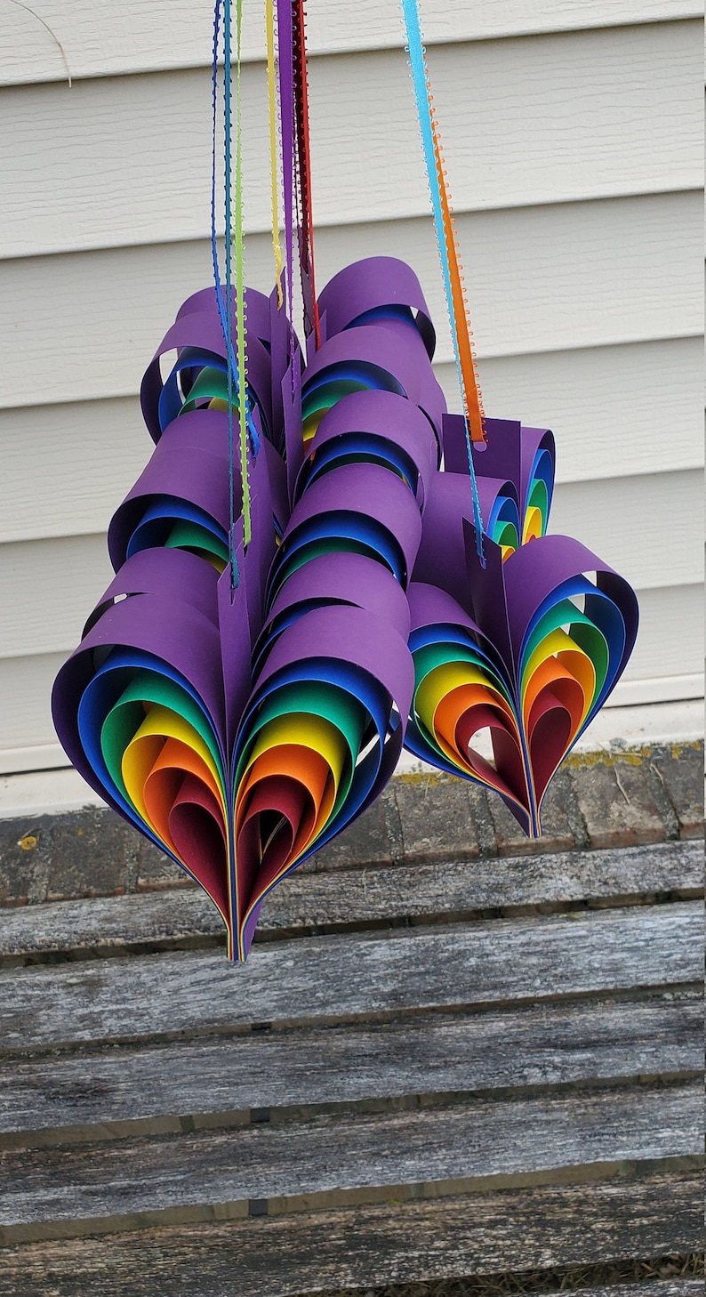 May include: A set of three rainbow heart-shaped paper ornaments hanging from strings. The hearts are made of multiple layers of paper in rainbow colors, with a purple outer layer. The hearts are hanging from a white wall.