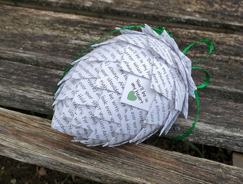 Pode incluir: Uma esfera de papel branco com fita verde, feita de notas de papel dobradas com mensagens escritas &agrave; m&atilde;o. A esfera &eacute; decorada com um pequeno cora&ccedil;&atilde;o verde e o texto "You'll be in my heart" escrito em uma etiqueta branca.