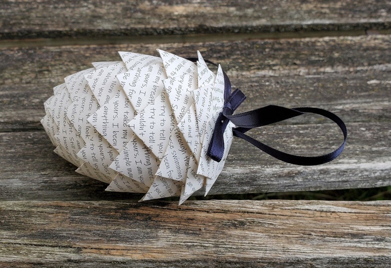 Pode incluir: Um enfeite de papel branco em forma de pinha com uma fita preta amarrada. O enfeite &eacute; decorado com texto escrito &agrave; m&atilde;o.