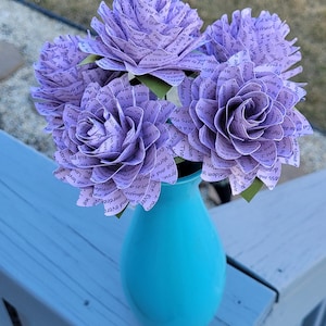 May include: A bouquet of purple paper flowers with text printed on them, arranged in a turquoise vase. The flowers are sitting on a light blue wooden surface.