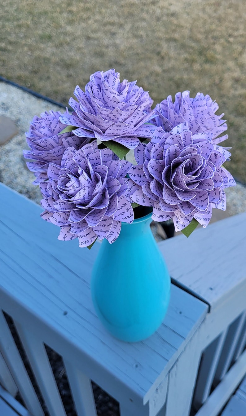 May include: A bouquet of purple paper flowers with text printed on them, arranged in a turquoise vase. The flowers are sitting on a light blue wooden surface.