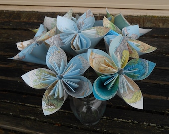 May include: A bouquet of origami flowers made from maps in shades of blue and beige. The flowers are arranged in a clear glass vase.