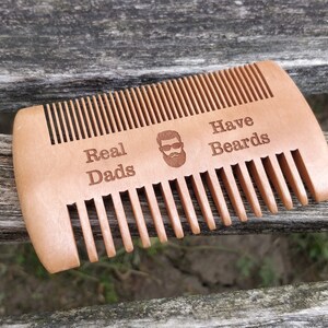 Peut inclure: Un peigne en bois avec le texte "Real Dads Have Beards" et une image de dessin anim&eacute; d'un homme avec une barbe et des lunettes de soleil.