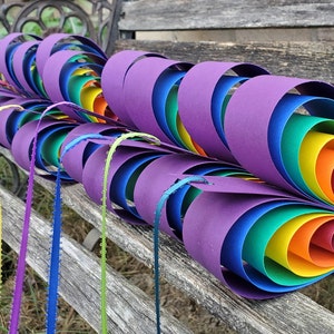 May include: A rainbow-colored paper craft in the shape of a heart, made of multiple layers of paper, with each layer a different color of the rainbow. The craft is sitting on a wooden bench.