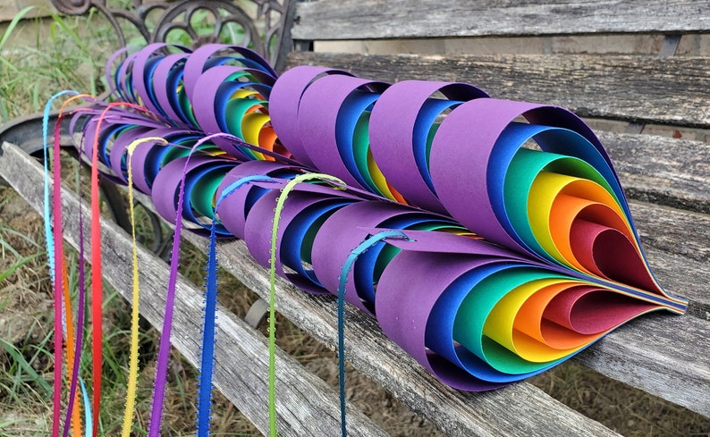 May include: A rainbow-colored paper craft in the shape of a heart, made of multiple layers of paper, with each layer a different color of the rainbow. The craft is sitting on a wooden bench.
