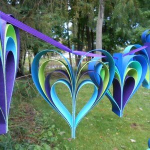 Pu&ograve; includere: Una ghirlanda di cuori di carta in tonalit&agrave; di blu, verde e viola. I cuori sono sovrapposti e piegati per creare un effetto tridimensionale. La ghirlanda &egrave; infilata su un nastro viola.