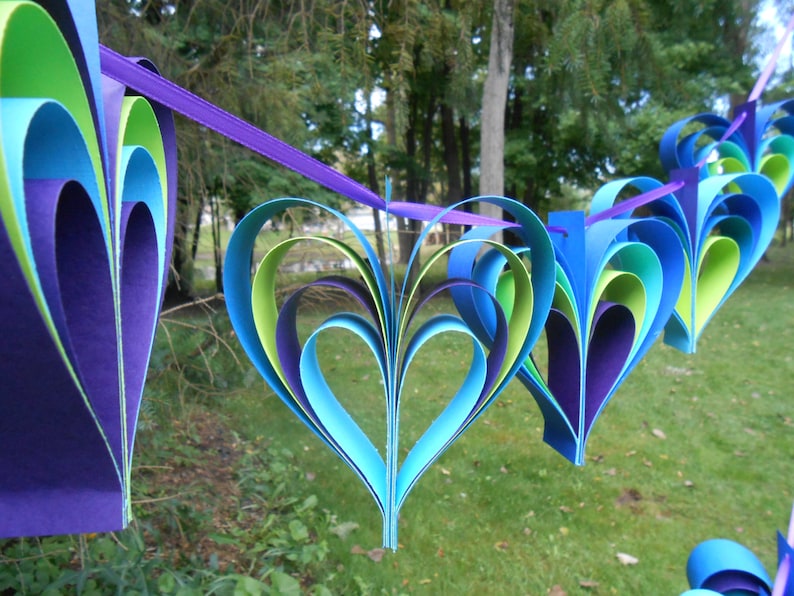 Pu&ograve; includere: Una ghirlanda di cuori di carta in tonalit&agrave; di blu, verde e viola. I cuori sono sovrapposti e piegati per creare un effetto tridimensionale. La ghirlanda &egrave; infilata su un nastro viola.