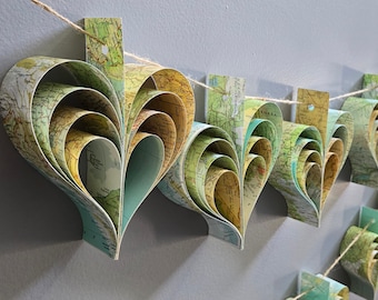 May include: A garland of paper hearts made from vintage maps. Each heart is layered and folded to create a three-dimensional effect. The garland is strung with twine and would be a unique and charming addition to a home decor or wedding.