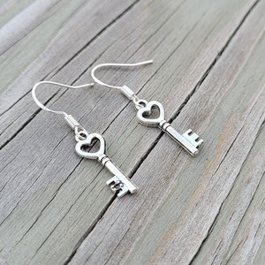 May include: A pair of silver key-shaped earrings with heart-shaped keyholes. The earrings are hanging from a wooden surface.