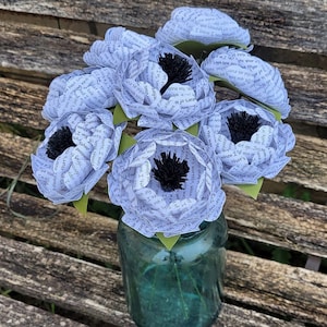 May include: A bouquet of six white paper flowers with black centers, arranged in a clear glass jar. The flowers are made from pages of a book, with the text visible on the petals.