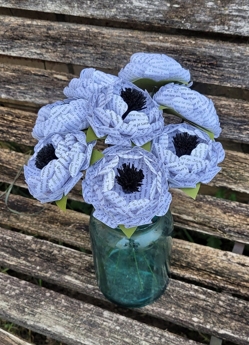 May include: A bouquet of six white paper flowers with black centers, arranged in a clear glass jar. The flowers are made from pages of a book, with the text visible on the petals.