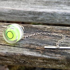Può includere: Un fermaglio per cravatta argentato con un livello a bolla rotondo trasparente. Il livello a bolla contiene un liquido verde e un anello bianco attorno al bordo.