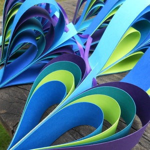 Pu&ograve; includere: Un cuore di carta artigianale realizzato con strisce di carta blu, verde e viola. Il cuore &egrave; piegato in un motivo a strati, creando un effetto tridimensionale.