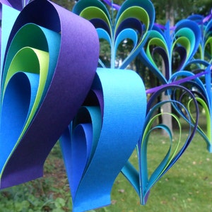 Pu&ograve; includere: Una ghirlanda di cuori di carta in tonalit&agrave; di blu, verde e viola. I cuori sono sovrapposti e piegati per creare un effetto tridimensionale.