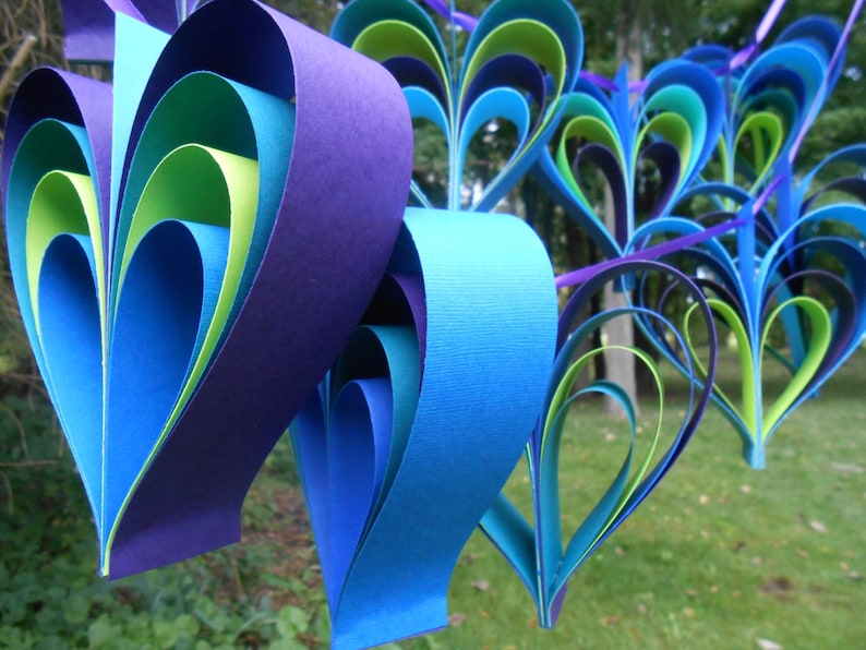 Pu&ograve; includere: Una ghirlanda di cuori di carta in tonalit&agrave; di blu, verde e viola. I cuori sono sovrapposti e piegati per creare un effetto tridimensionale.