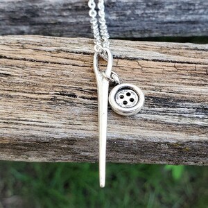 May include: A silver necklace with a needle and button charm. The needle is long and thin, and the button is small and round. The necklace is on a wooden surface.