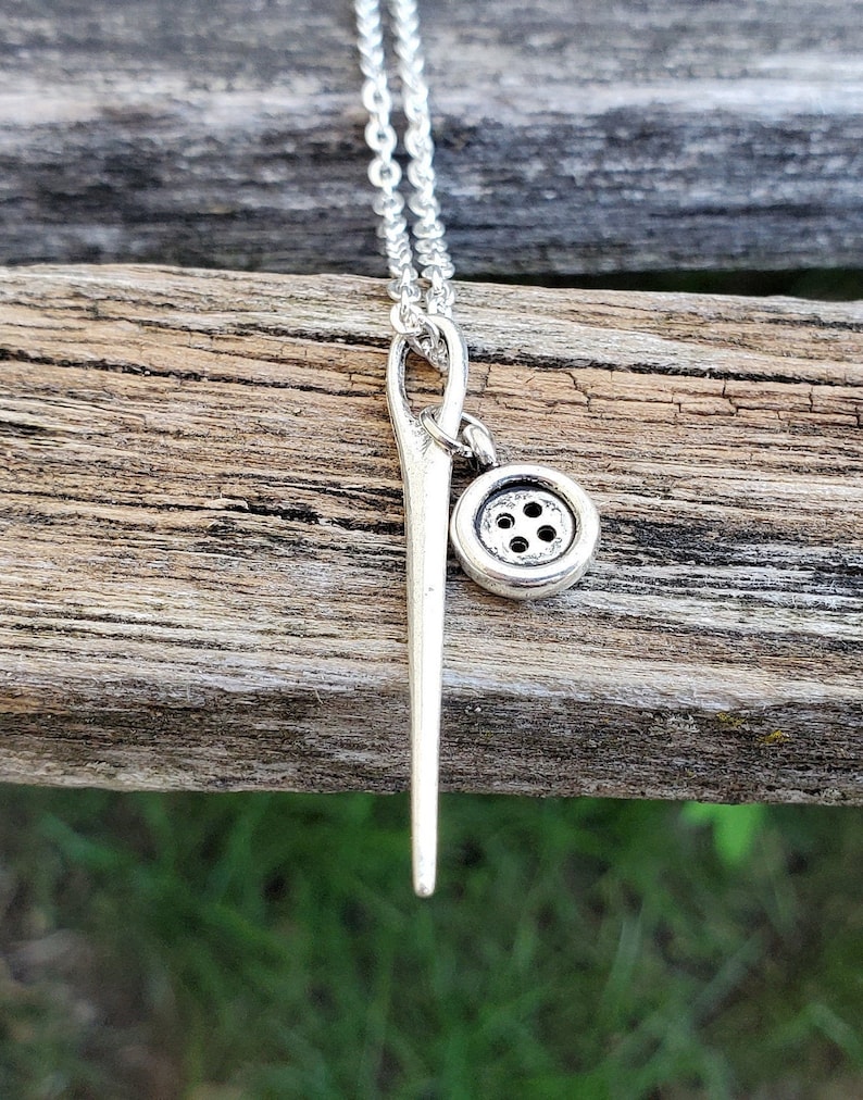 May include: A silver necklace with a needle and button charm. The needle is long and thin, and the button is small and round. The necklace is on a wooden surface.
