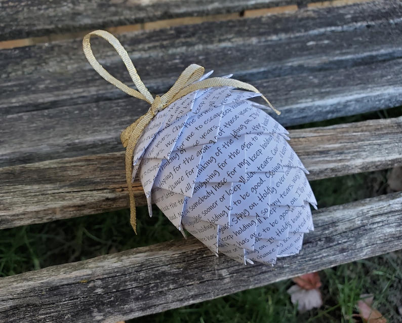 Pode incluir: Um enfeite de papel branco em forma de pinha com uma fita dourada. O enfeite &eacute; coberto com texto escrito &agrave; m&atilde;o, incluindo as palavras "escape", "hoping for", "chance for", "keep holding", "to be good", "holding on", "my tears", "every time", "keep tears", "good on", "choice", "holding", "so good".