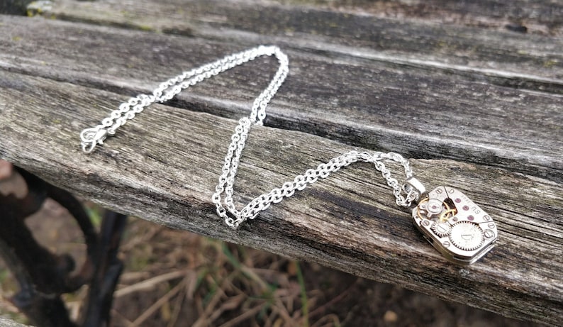 May include: A silver chain necklace with a silver pendant featuring a detailed clockwork design. The pendant is rectangular and has visible gears and other clock parts.