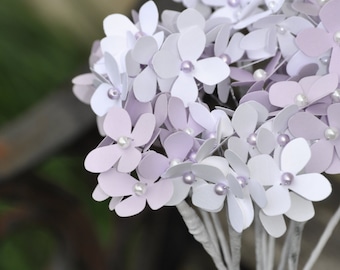 May include: A bouquet of paper flowers in a clear glass vase. The flowers are white and lavender with pearl centers. The stems are white and the vase is partially visible.