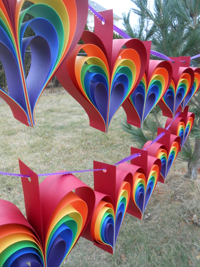 May include: A rainbow paper heart garland with red, orange, yellow, green, blue, indigo, and violet paper hearts strung together with purple string.