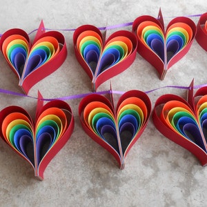 May include: A garland of ten rainbow paper hearts strung together with purple ribbon. Each heart is made of layered paper circles in the colors of the rainbow.