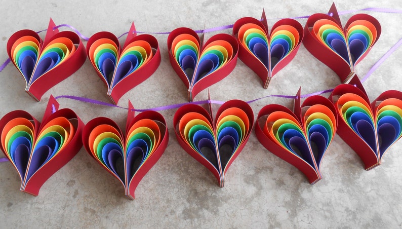 May include: A garland of ten rainbow paper hearts strung together with purple ribbon. Each heart is made of layered paper circles in the colors of the rainbow.