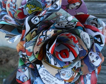 Puede incluir: Un ramo de rosas hecho de páginas de cómics. Las rosas son rojas, azules y amarillas. Las páginas de cómics presentan personajes y diálogos del universo DC Comics. El texto en las páginas incluye "Harley no corre! Ella se ha enfrentado a eso", "Holtee", "Dáselas a él", y "Contrato abierto sobre Quinn! Un tipo la quiere muerta!"