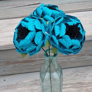 May include: Three turquoise paper flowers with black centers are arranged in a clear glass bottle. The flowers are made of paper with text printed on them. The bottle is sitting on a wooden surface.