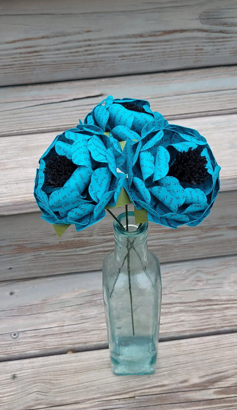 May include: Three turquoise paper flowers with black centers are arranged in a clear glass bottle. The flowers are made of paper with text printed on them. The bottle is sitting on a wooden surface.