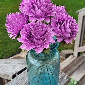 May include: A bouquet of five purple paper roses in a blue glass jar. The roses are made of paper and have words written on them. The words are in black ink and are visible on the petals.