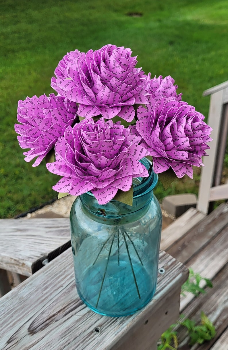 May include: A bouquet of five purple paper roses in a blue glass jar. The roses are made of paper and have words written on them. The words are in black ink and are visible on the petals.