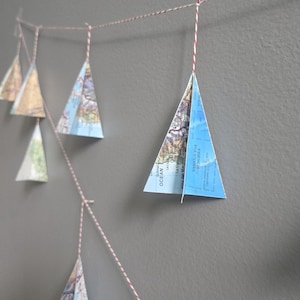 May include: A decorative garland featuring multiple triangular paper ornaments crafted from vintage maps. Each ornament is suspended from a red and white baker's twine string. The map segments display blue ocean areas and landmasses with text.