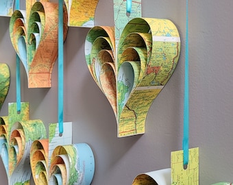 May include: A collection of seven heart-shaped paper ornaments made from rolled up maps, hanging from blue ribbons against a gray wall.
