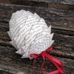 Pode incluir: Um ornamento de papel branco em forma de pinha, dobrado de uma p&aacute;gina de livro com texto preto. O ornamento est&aacute; amarrado com uma fita vermelha.
