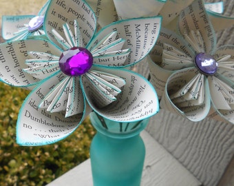 Puede incluir: Un ramo de flores de papel hechas a mano en un jarrón turquesa. Las flores están hechas de páginas de libros, con bordes turquesa y centros de gemas moradas. El jarrón es de color turquesa esmerilado.