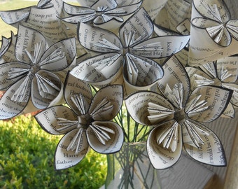Puede incluir: Un ramo de flores de papel hechas con páginas de libros. Las flores son blancas con detalles negros y están dispuestas en un jarrón de vidrio transparente. Las flores están enfocadas, mientras que el fondo está desenfocado.