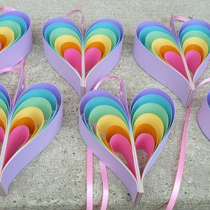 K&ouml;nnte beinhalten: Acht regenbogenfarbene, mehrschichtige Papierherzen mit rosa Schleifenb&auml;ndern. Die Herzen bestehen aus mehreren Papierschichten in den Farben Blau, Gr&uuml;n, Gelb, Orange und Rosa.