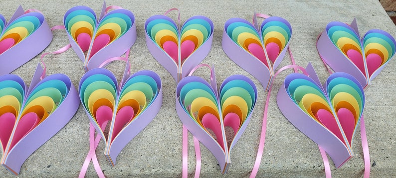 K&ouml;nnte beinhalten: Acht regenbogenfarbene, mehrschichtige Papierherzen mit rosa Schleifenb&auml;ndern. Die Herzen bestehen aus mehreren Papierschichten in den Farben Blau, Gr&uuml;n, Gelb, Orange und Rosa.