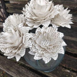 May include: A bouquet of white paper flowers made from book pages. The flowers are arranged in a clear glass vase. The vase is sitting on a wooden bench.