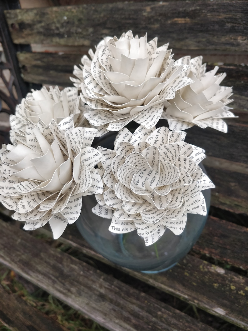 May include: A bouquet of white paper flowers made from book pages. The flowers are arranged in a clear glass vase. The vase is sitting on a wooden bench.