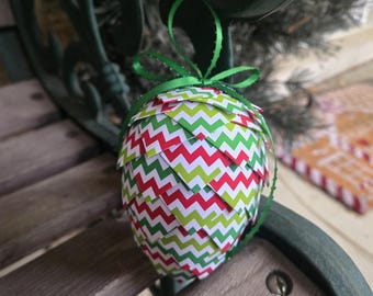 Könnte beinhalten: Handgefertigter Weihnachtsschmuck in Form eines Tannenzapfens. Der Schmuck besteht aus geschichtetem Papier mit einem roten, grünen und weißen Zickzackmuster. Ein grünes Band ist zum Aufhängen befestigt. Der Schmuck wird auf einer Holzoberfläche präsentiert.