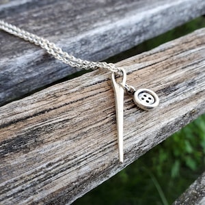 May include: A silver necklace with a needle and button charm. The necklace is on a wooden surface.