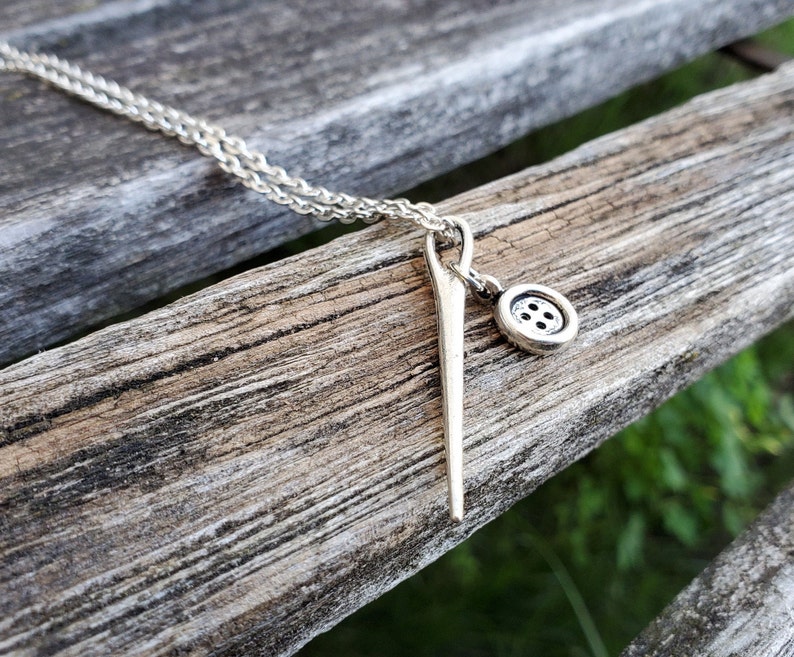 May include: A silver necklace with a needle and button charm. The necklace is on a wooden surface.
